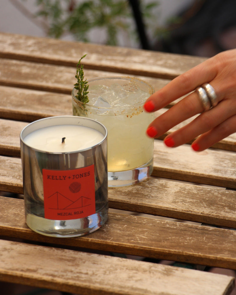 A woman's hand reaching for a mezcal cocktail next to a Mezcal Roja candle outside on a patio of a mezcal bar